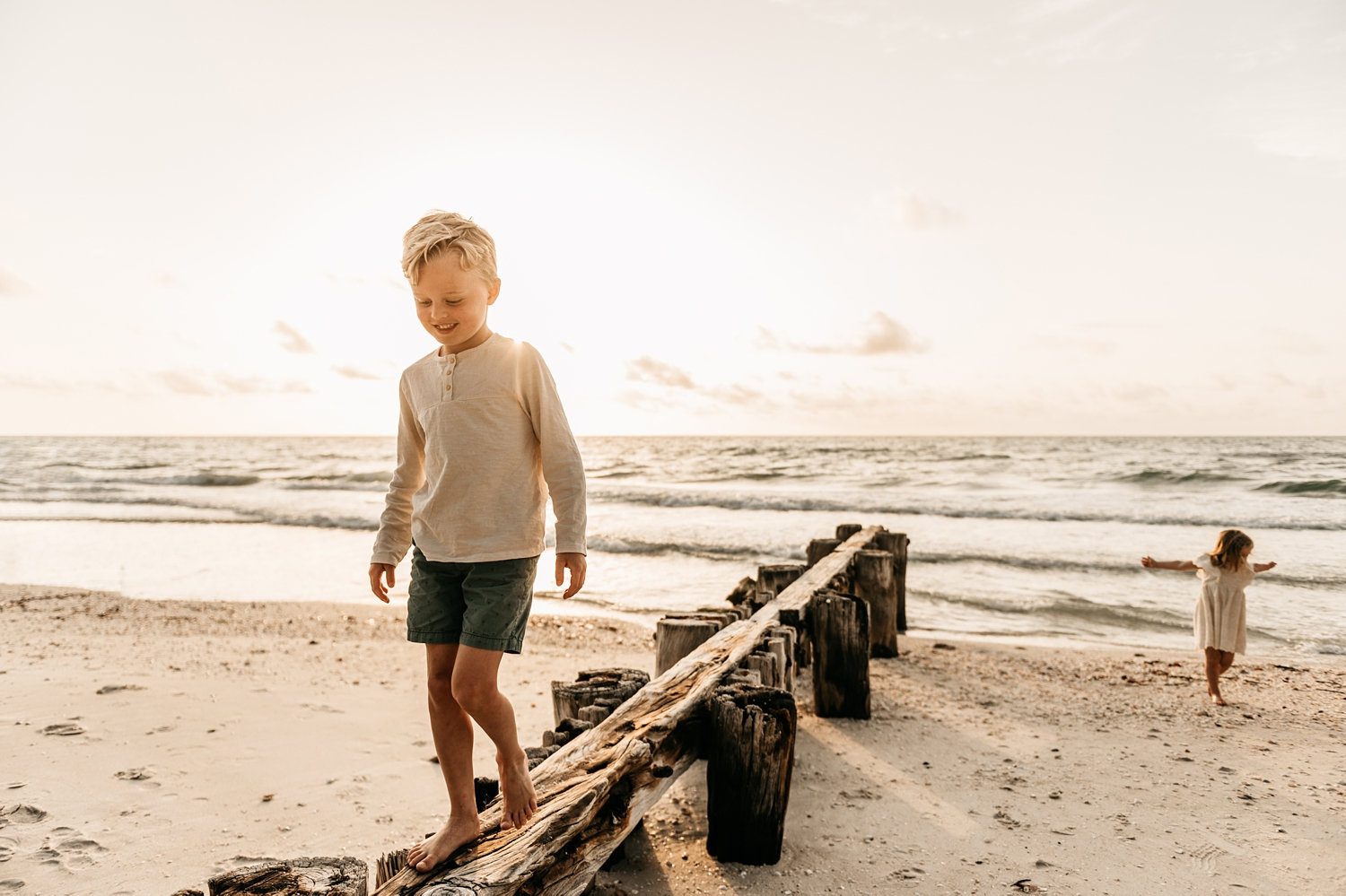 blonde boy walking on beach pilings with little sister in background, Ryaphotos