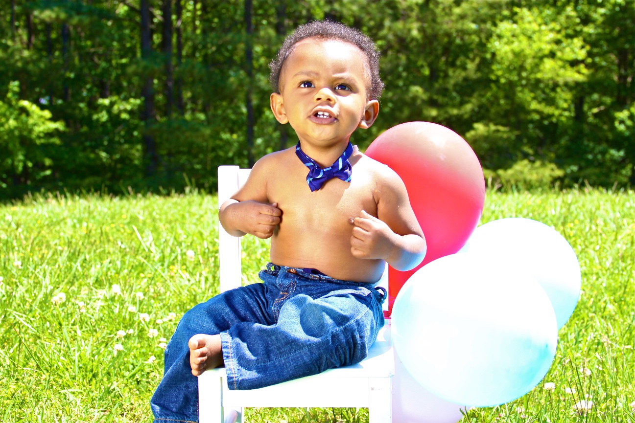 Young boy sitting on a chair outdoors with colorful balloons behind him.