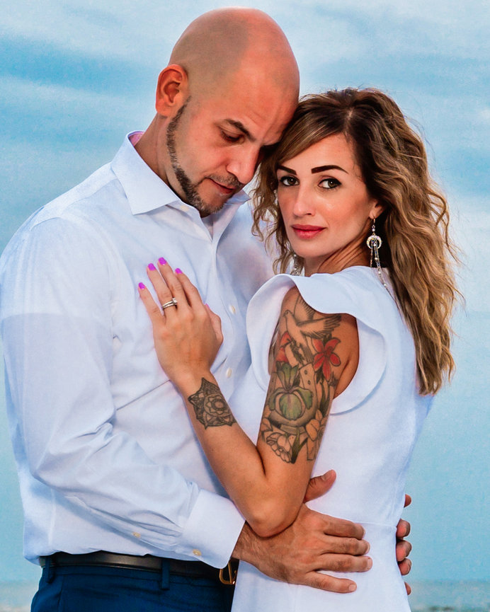 Couple embracing outdoors, showcasing tattoos and dress attire against a cloudy sky.