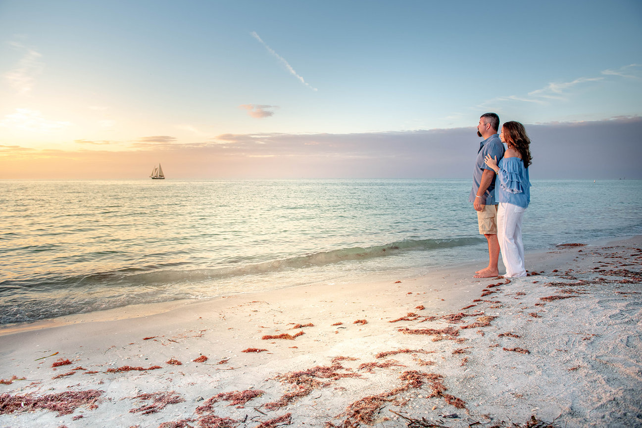Couple standing on a beach at sunset, gazing at the ocean with a sailing boat in the distance.