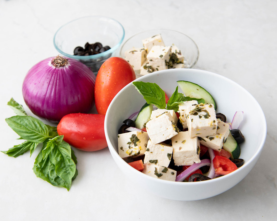 Fresh salad with tofu, tomatoes, cucumber, red onion, olives, and basil on a marble surface.