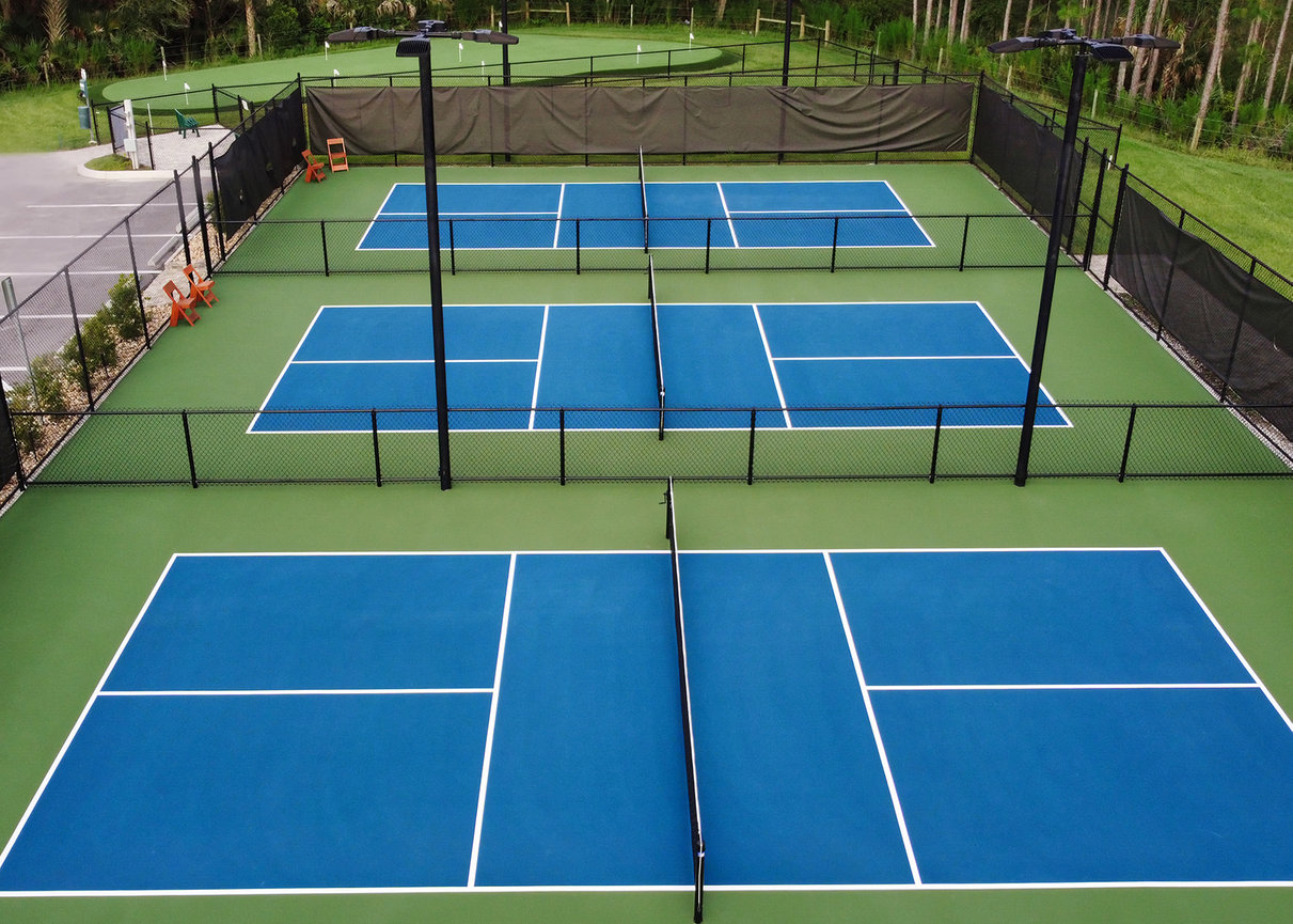 Aerial view of multiple tennis courts with blue surfaces and surrounding fencing in a green park setting.