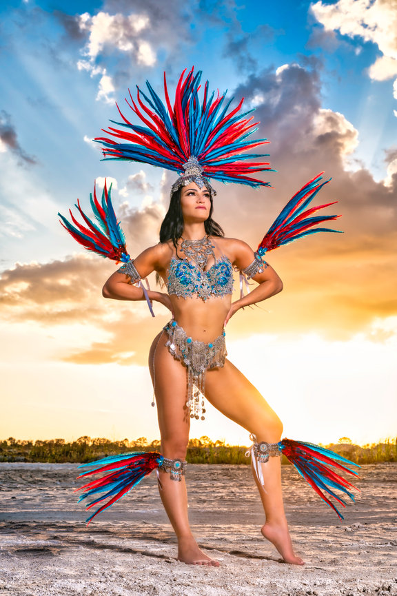 Woman in vibrant feathered costume poses confidently against a sunset backdrop.