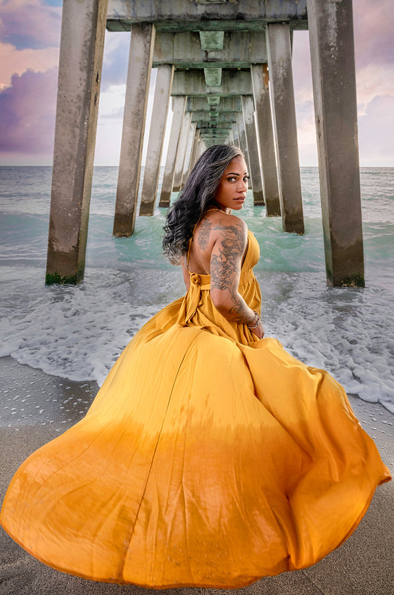 A woman in a yellow dress stands under a pier, with ocean waves and a cloudy sky in the background.