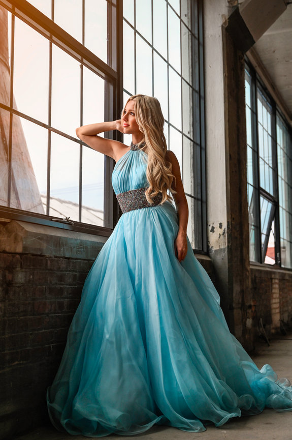 Woman in a flowing blue gown poses by a large window, showcasing elegant fabric and a serene expression.
