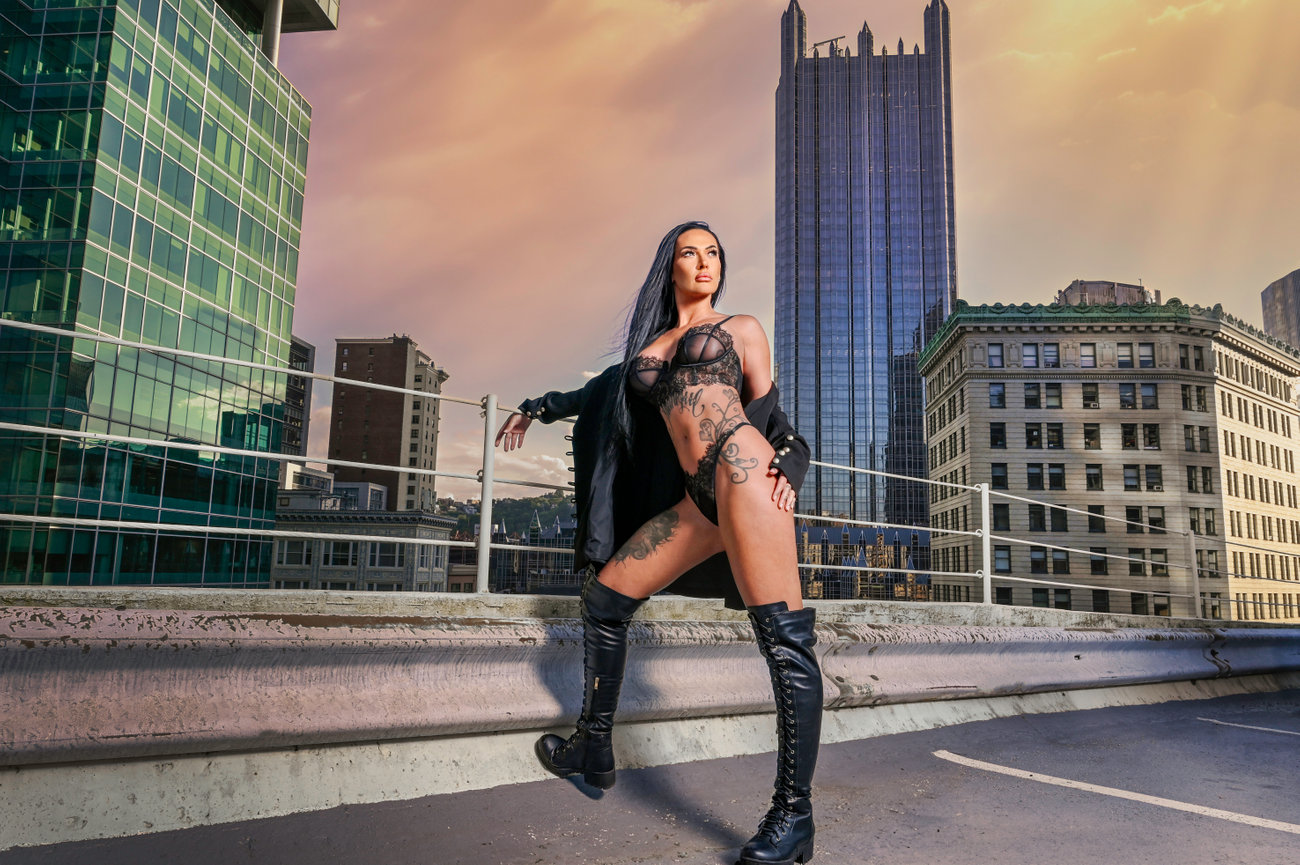 Luxury boudoir client posing on a rooftop at sunset, wearing lingerie and thigh-high boots with a cityscape view.