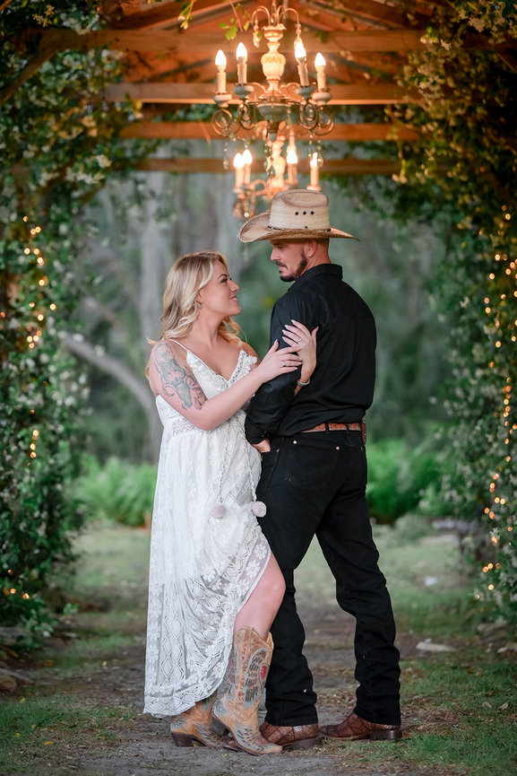 A couple embraces under a decorated archway with lights, wearing wedding attire and cowboy boots.