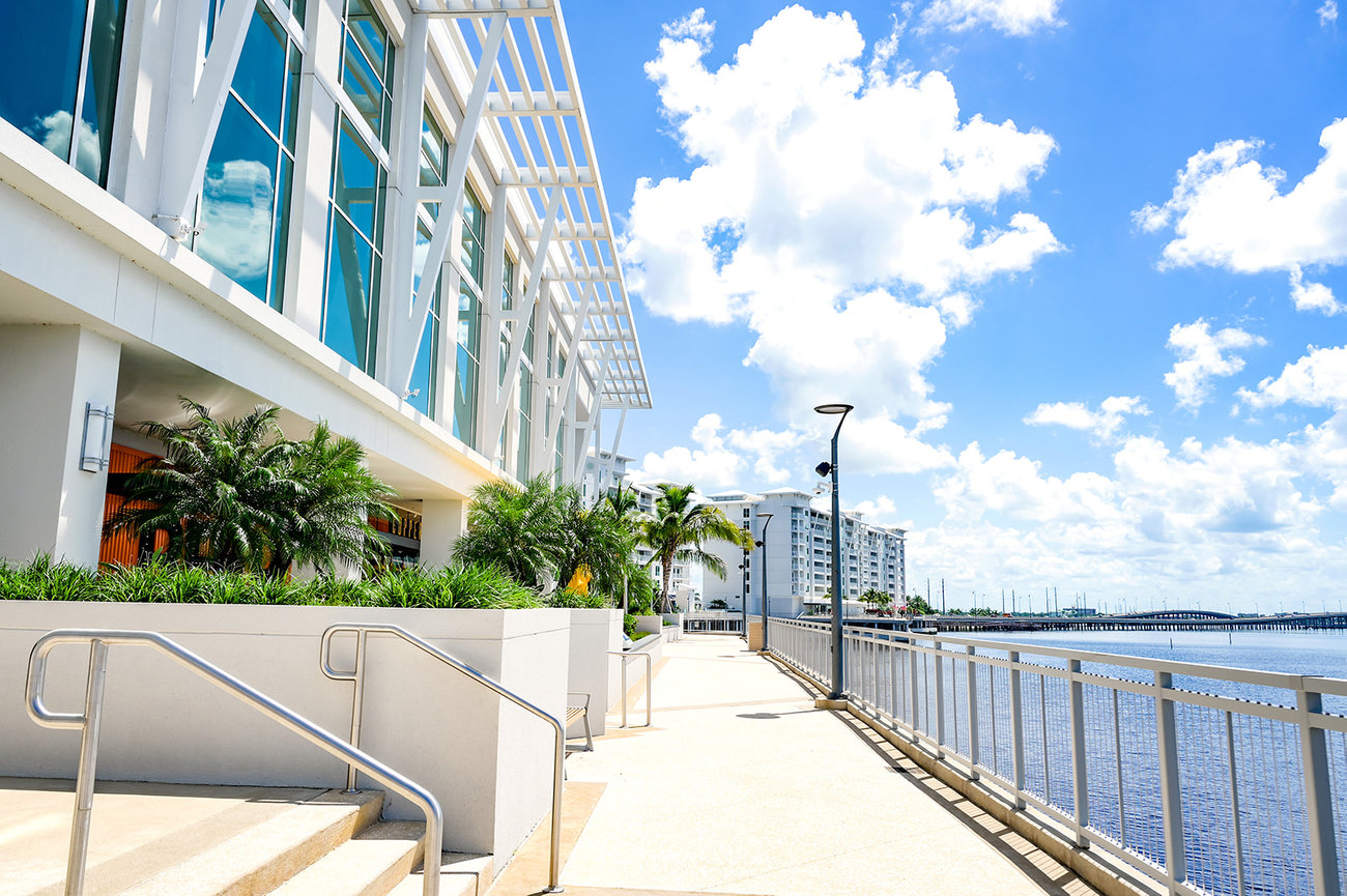 Modern waterfront building with large windows, palm trees, and a walkway along the water under a blue sky.