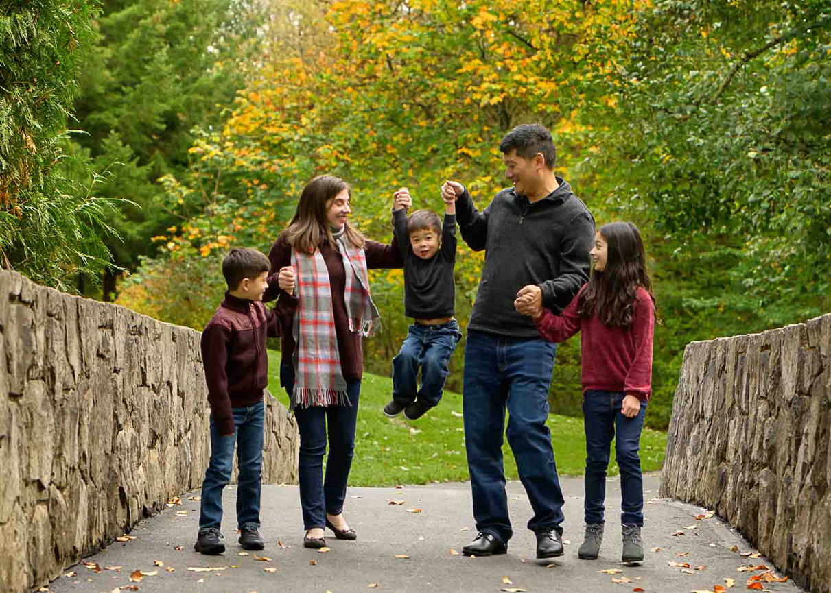 Family of five looking at each other and lifting the youngest up in the air as they walk across a bridge in the fall