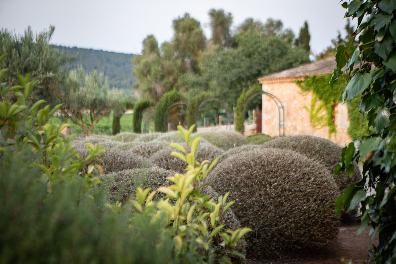 Garden with trimmed bushes and greenery, stone building in the background, and an archway path amidst lush vegetation. Mallorca