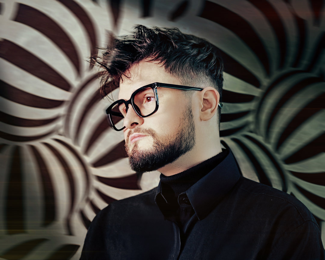 Young man with glasses and a beard, posing in front of a patterned background.