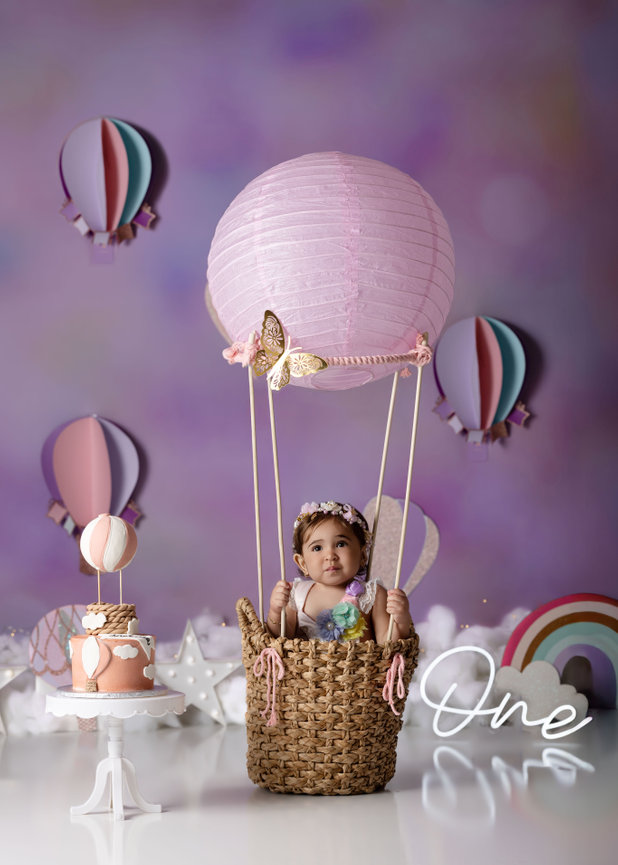 Baby in a basket balloon setup with a cake and decor, celebrating first birthday.
