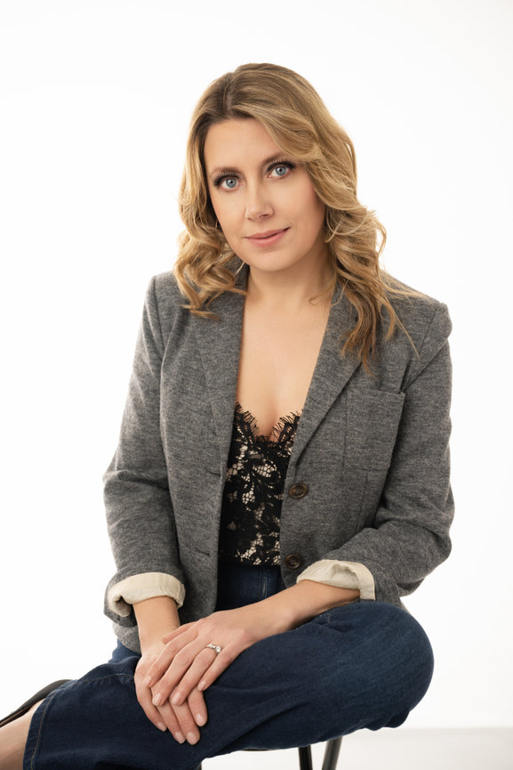 Woman with blonde hair in a gray blazer and black lace top sitting on a stool, looking at the camera.