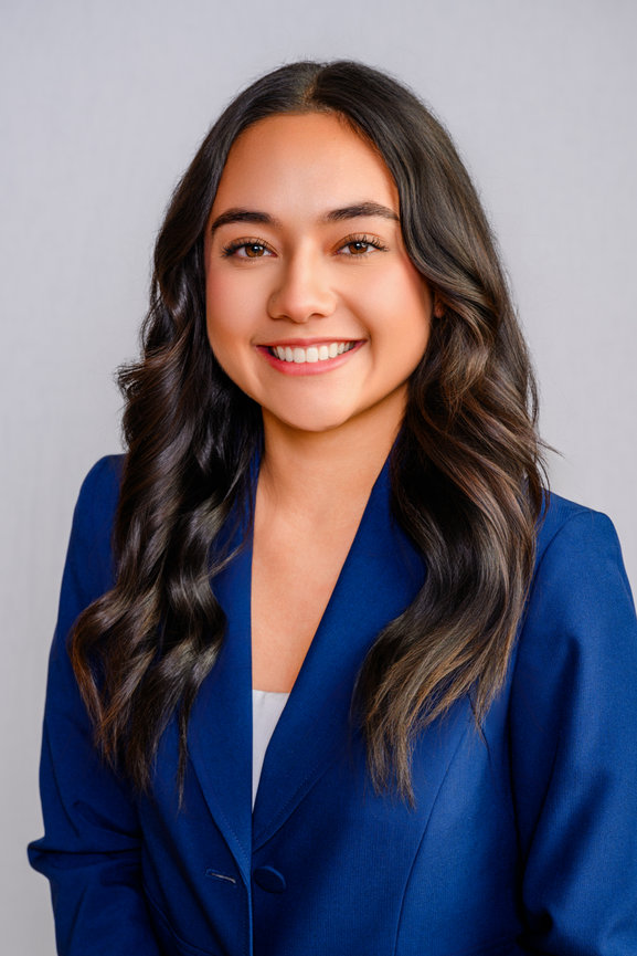 Smiling person with long wavy hair wearing a blue blazer against a neutral background.
