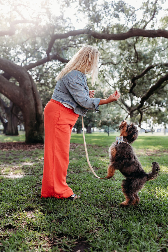 A woman in orange pants teaching a dog to stand on hind legs in a grassy park.