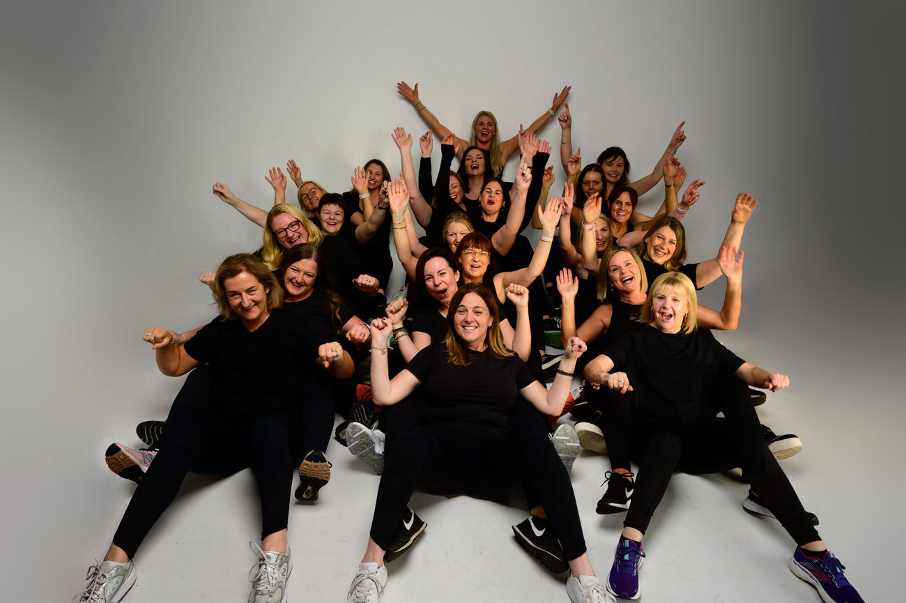 Group of women in black outfits, sitting and cheering energetically in a studio setting.