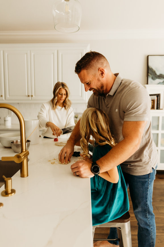 Family baking together in a bright kitchen, with a father helping a child at the counter while a woman prepares ingredients.
