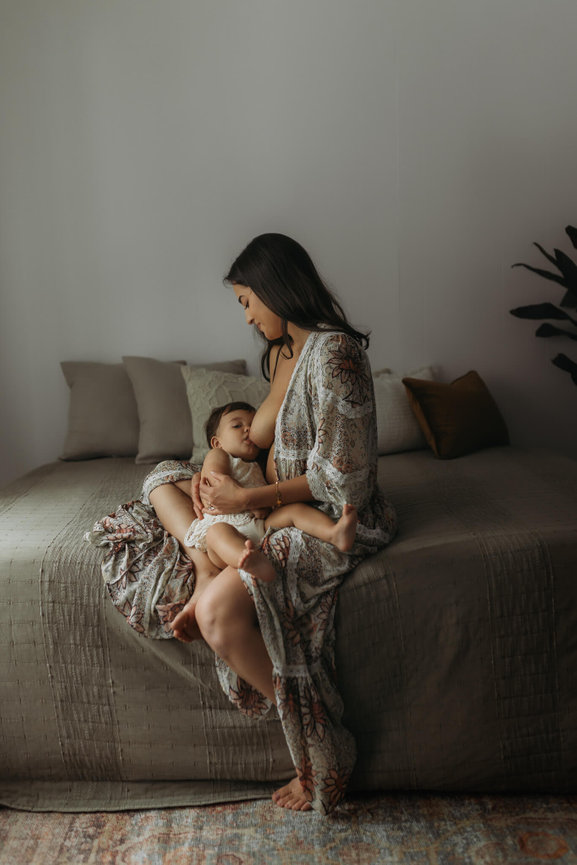 A mother breastfeeds her baby on the edge of a bed during a baby photography session in Mendham, New Jersey.