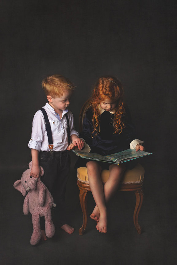 Redheaded siblings read a book together. The little boy holds a pink elephant lovey.