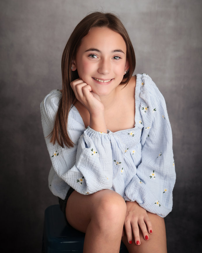 Smiling girl with long hair wearing a light blue, floral-patterned blouse, sitting against a gray backdrop.
