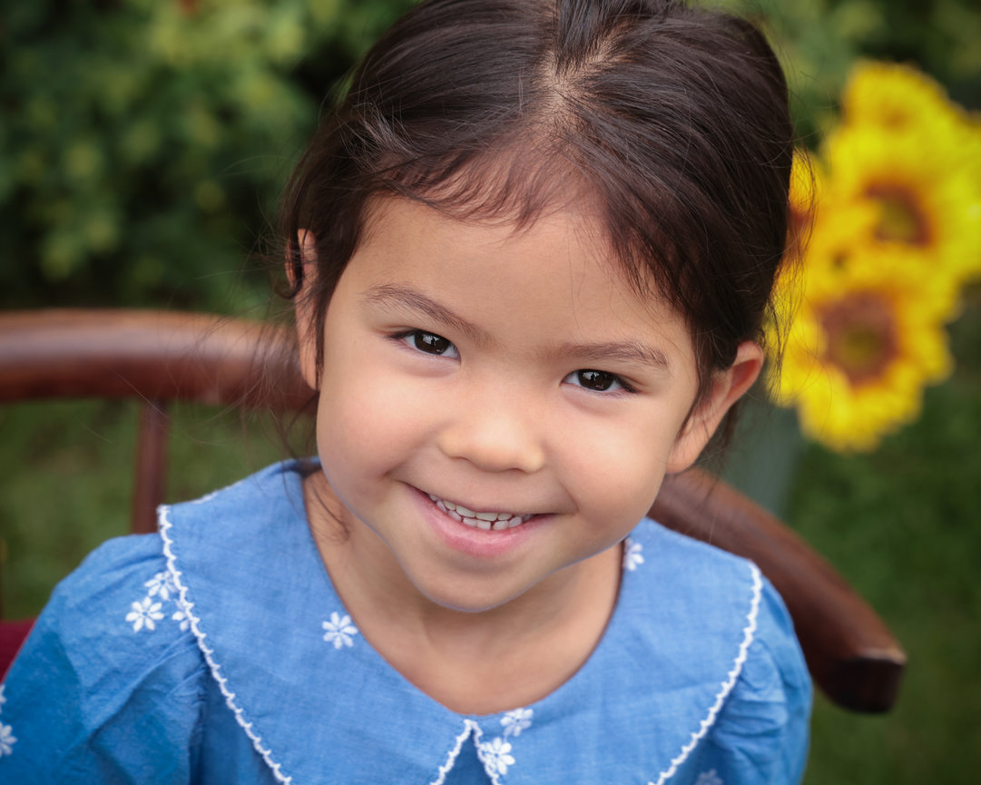 Young child in a blue dress smiling outdoors with sunflowers in the background.