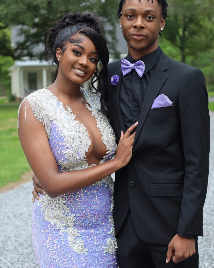A couple poses in formal attire outdoors, with the woman in a purple sequined dress and the man in a black suit with a purple bow tie.