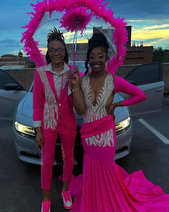 Two people in vibrant pink outfits stand in front of a car, one holding a feathered umbrella under a sunset sky.