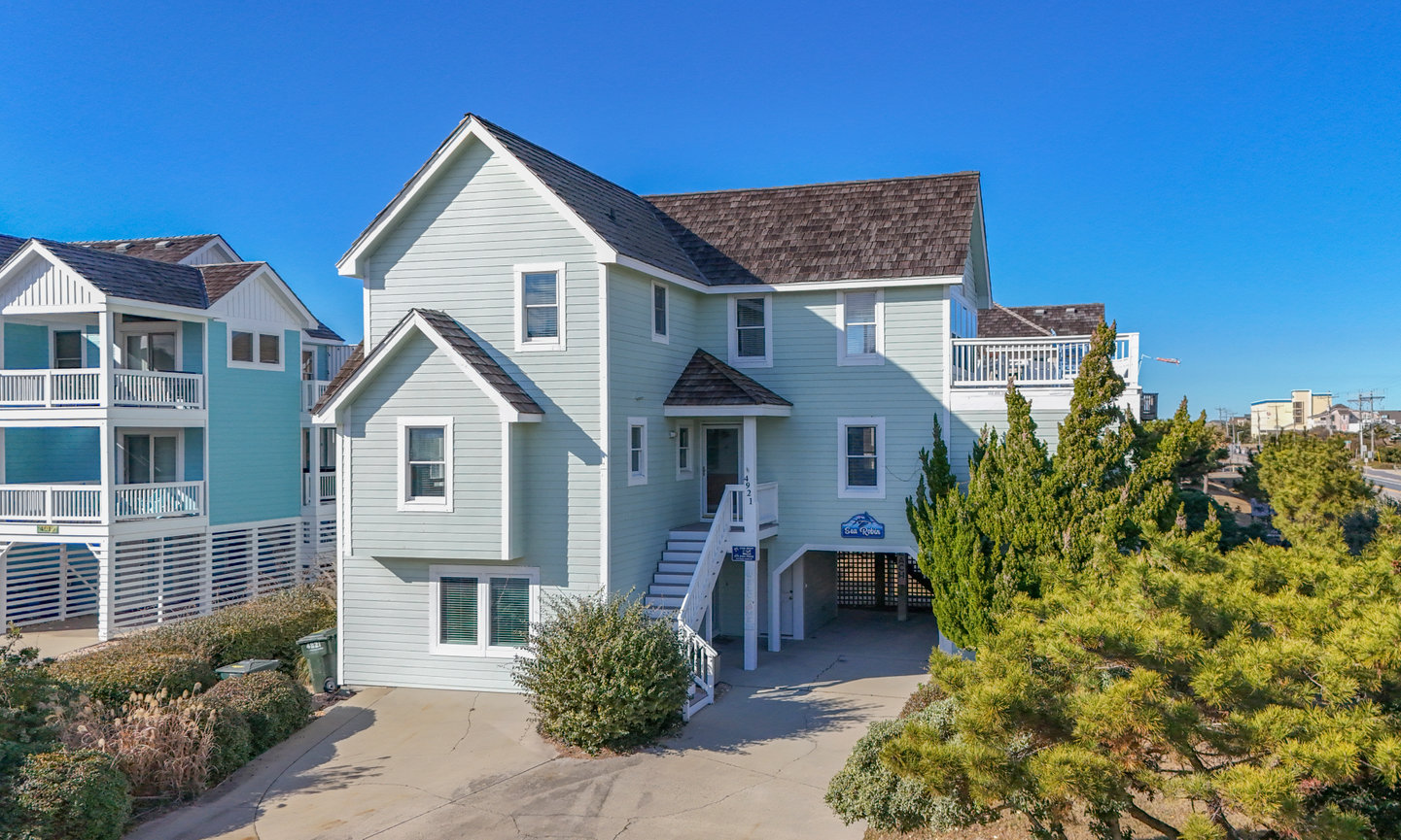 Light blue coastal house with a steep roof, surrounded by greenery, under a clear blue sky.