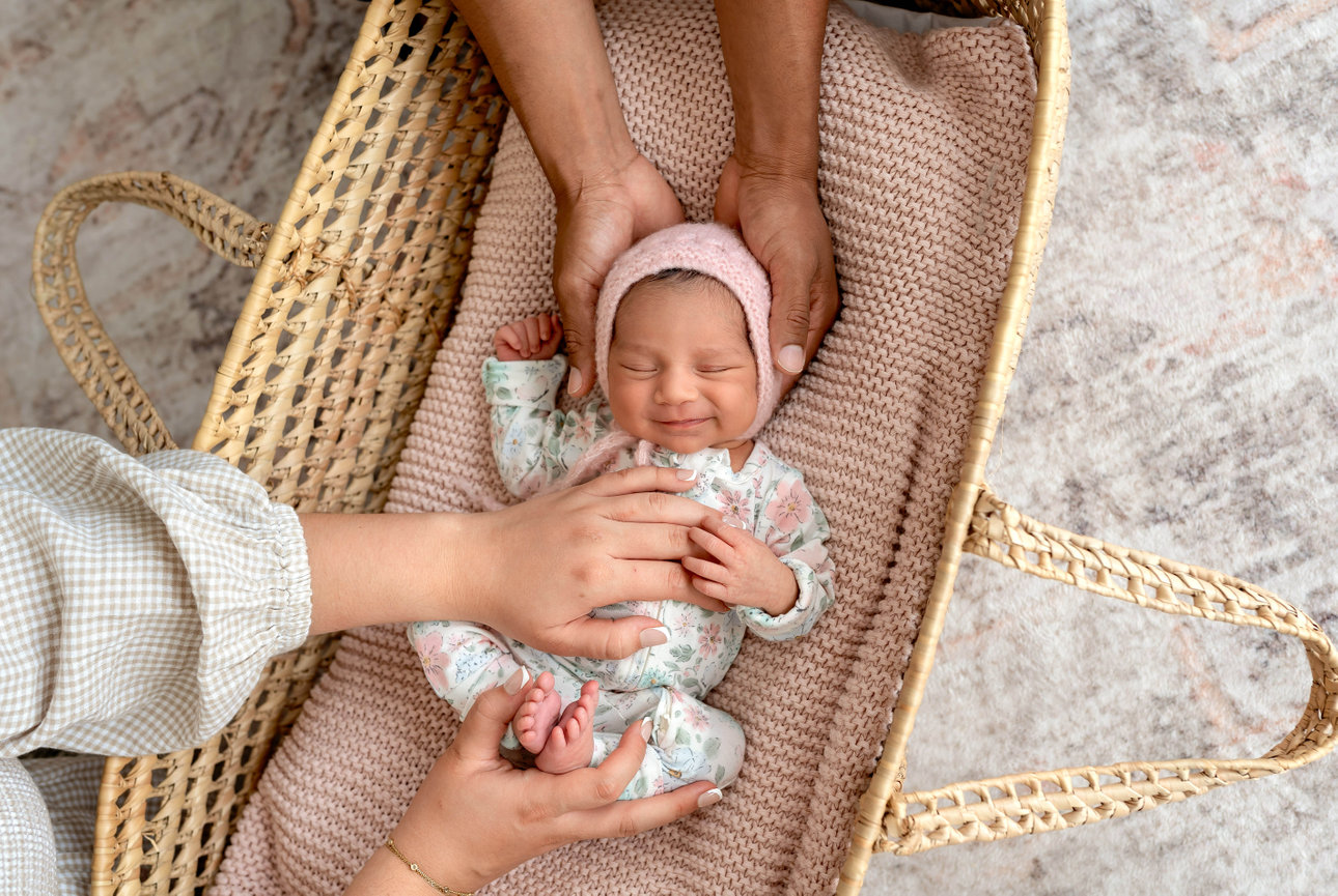 Newborn baby in a wicker bassinet, gently held by adult hands, wearing a pink bonnet and floral onesie.
