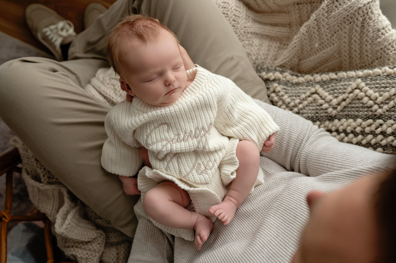 Sleeping baby in a cream knit outfit being held on a cozy blanket.