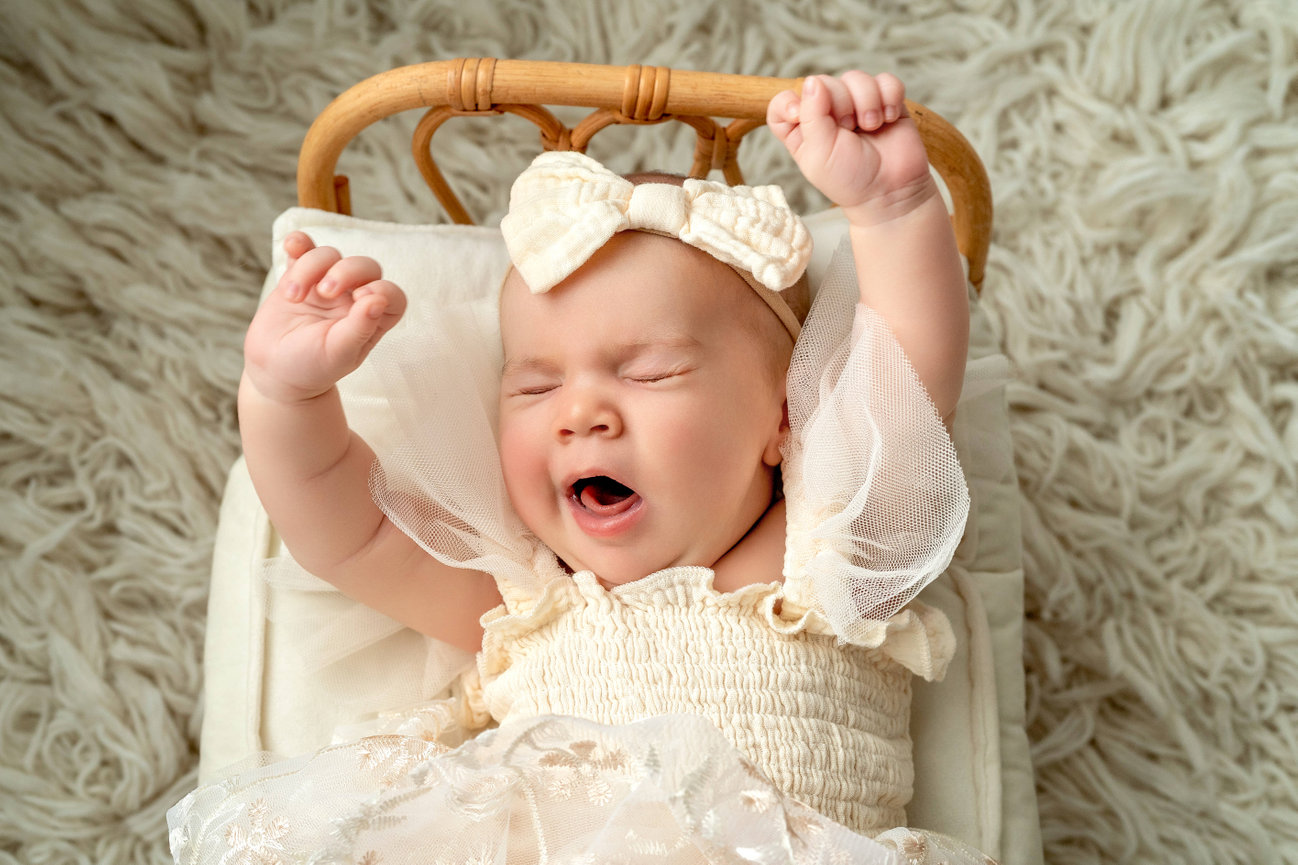 Yawning baby girl in a cream dress and bow, lying in a wicker cradle with arms raised