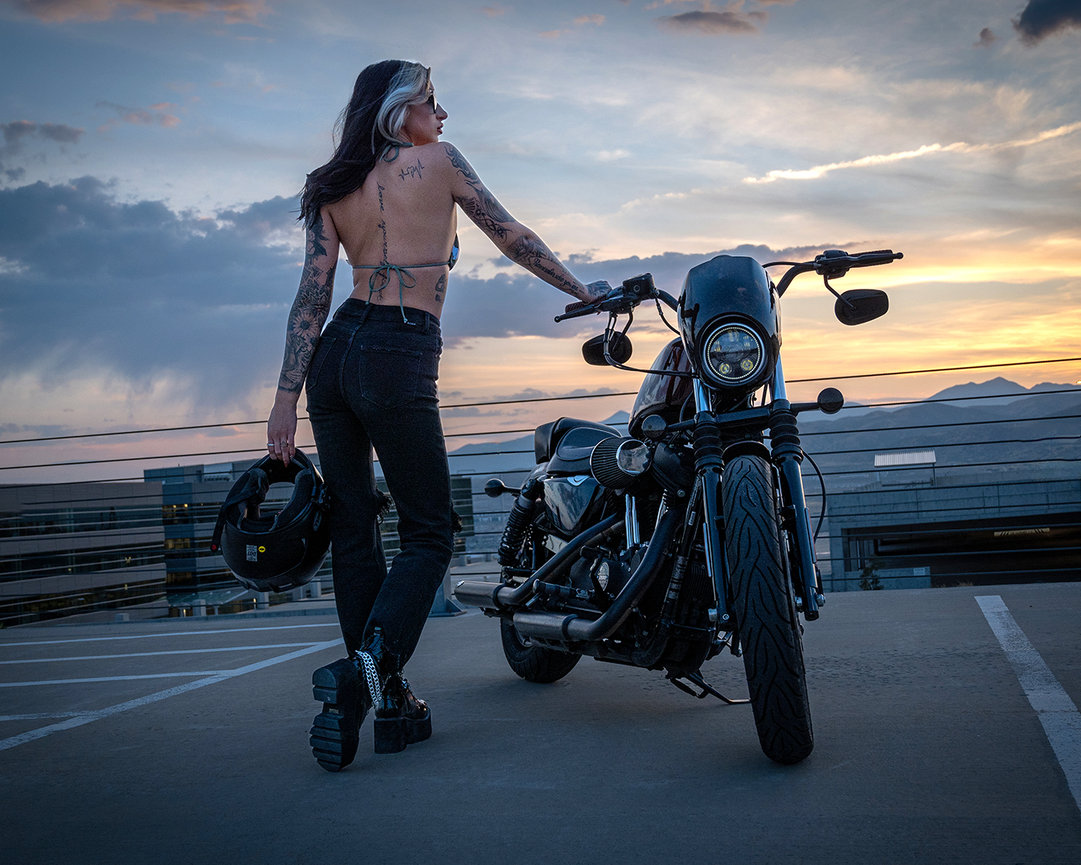 A tattooed woman standing next to a custom Harley Sportster motorcycle at sunset in Lehi, Utah, holding her helmet and looking into the distance, captured by Tre Nelson Photography.
