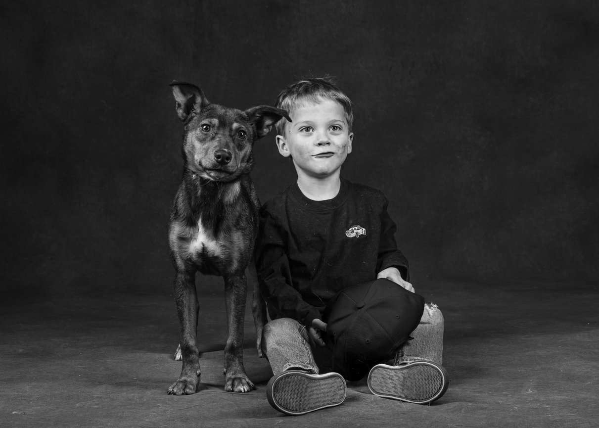 Black-and-white studio portrait of a young boy with smudged cheeks sitting beside a mixed-breed dog, both facing the camera with matching expressions.
