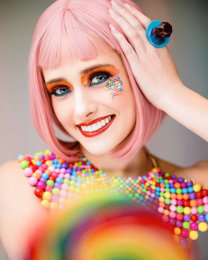 Woman with pink bob haircut wearing colorful candy-themed makeup and outfit, holding a lollipop.
