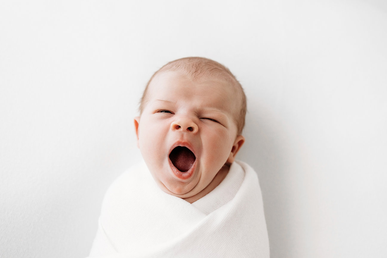 A baby wrapped in a white blanket yawning against a plain background.