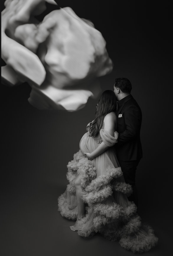 A couple in formal attire embraces, next to a large, abstract flower sculpture in grayscale.