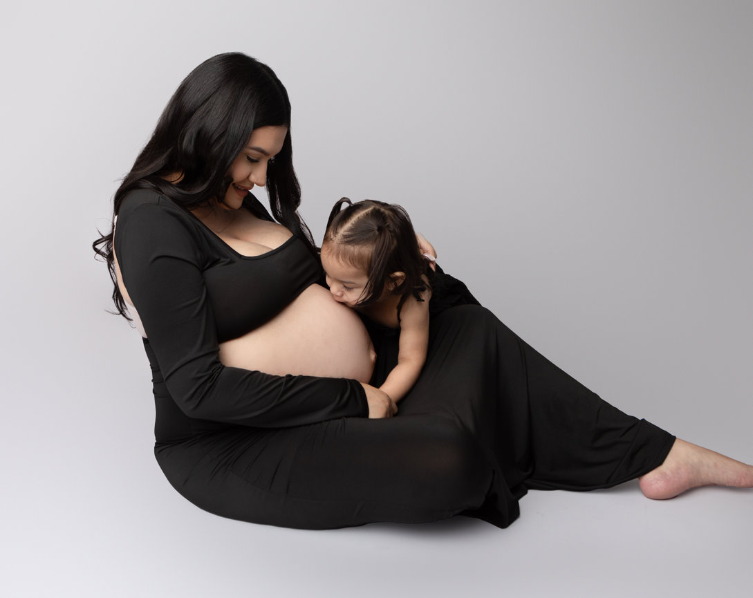 Pregnant woman in black dress sits while a young child kisses her belly against a soft gray background.