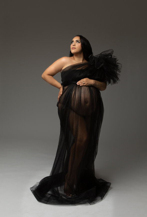 Pregnant woman in a sheer black gown posing confidently against a neutral background.