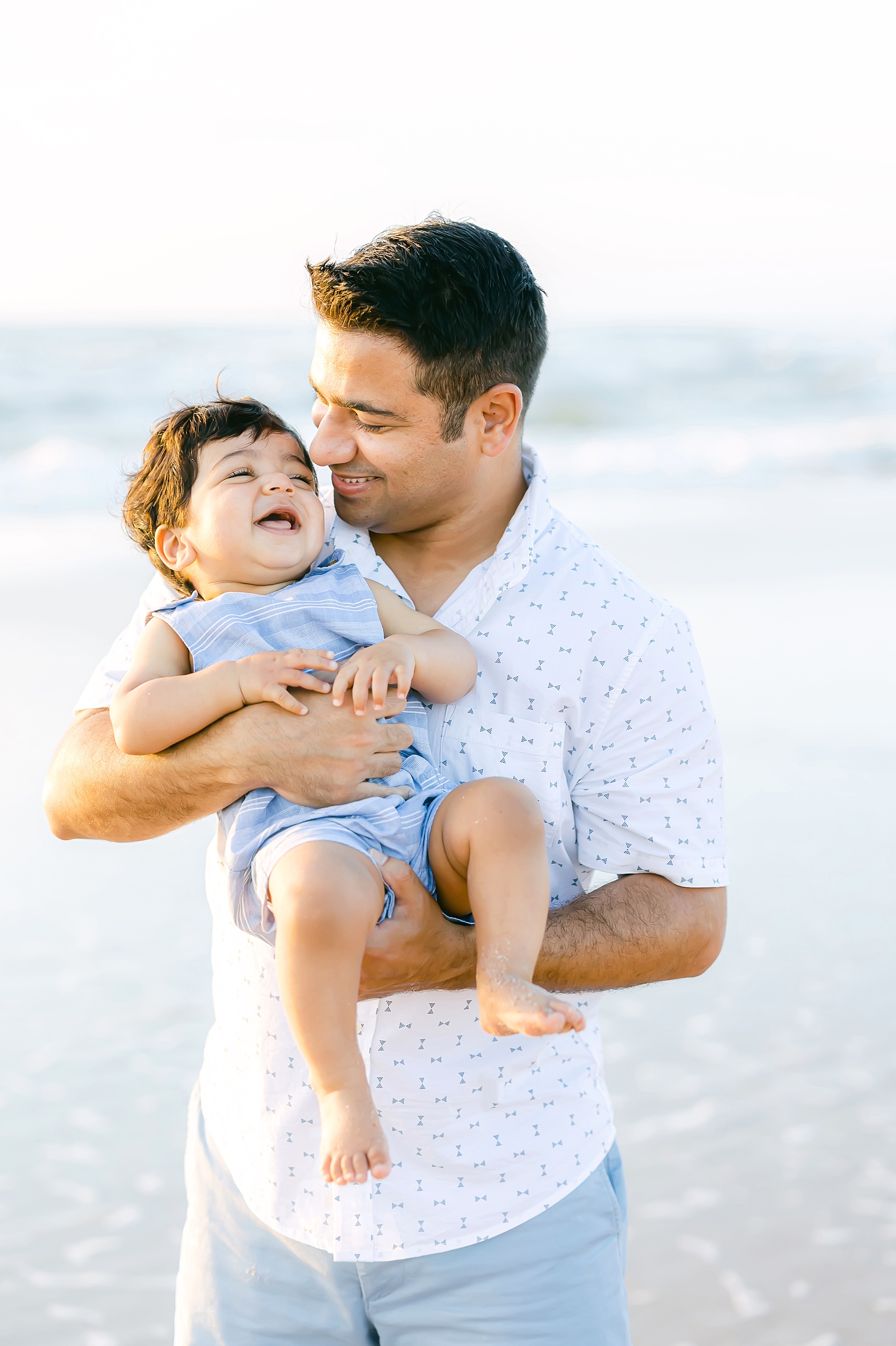 Dad holding baby laughing on the beach wearing blue and white
