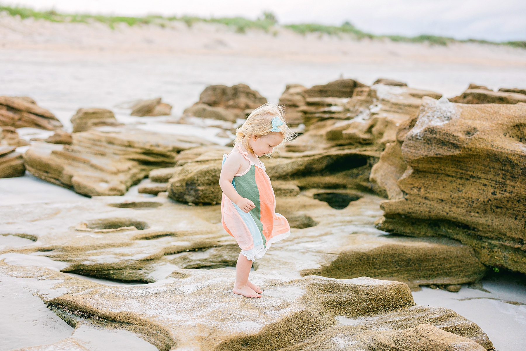 little girl in pastel striped dress standing on rocks on the beach