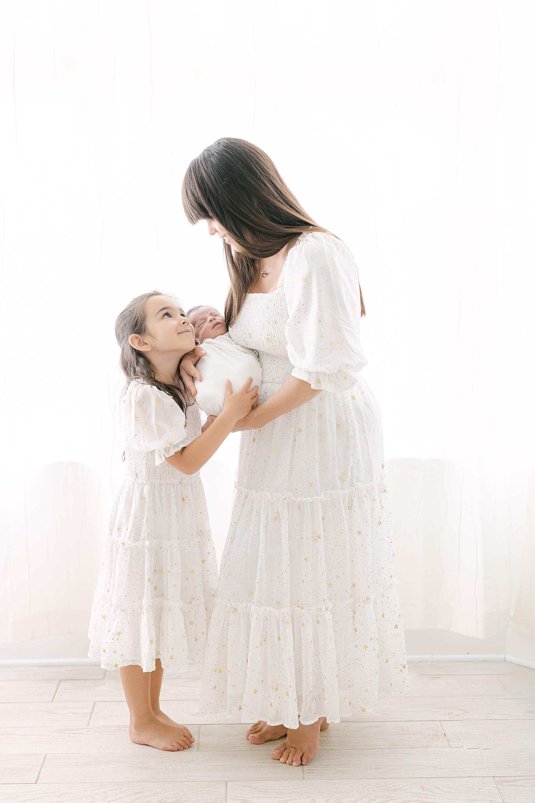 mother and daughter in white dresses holding baby in front of window light