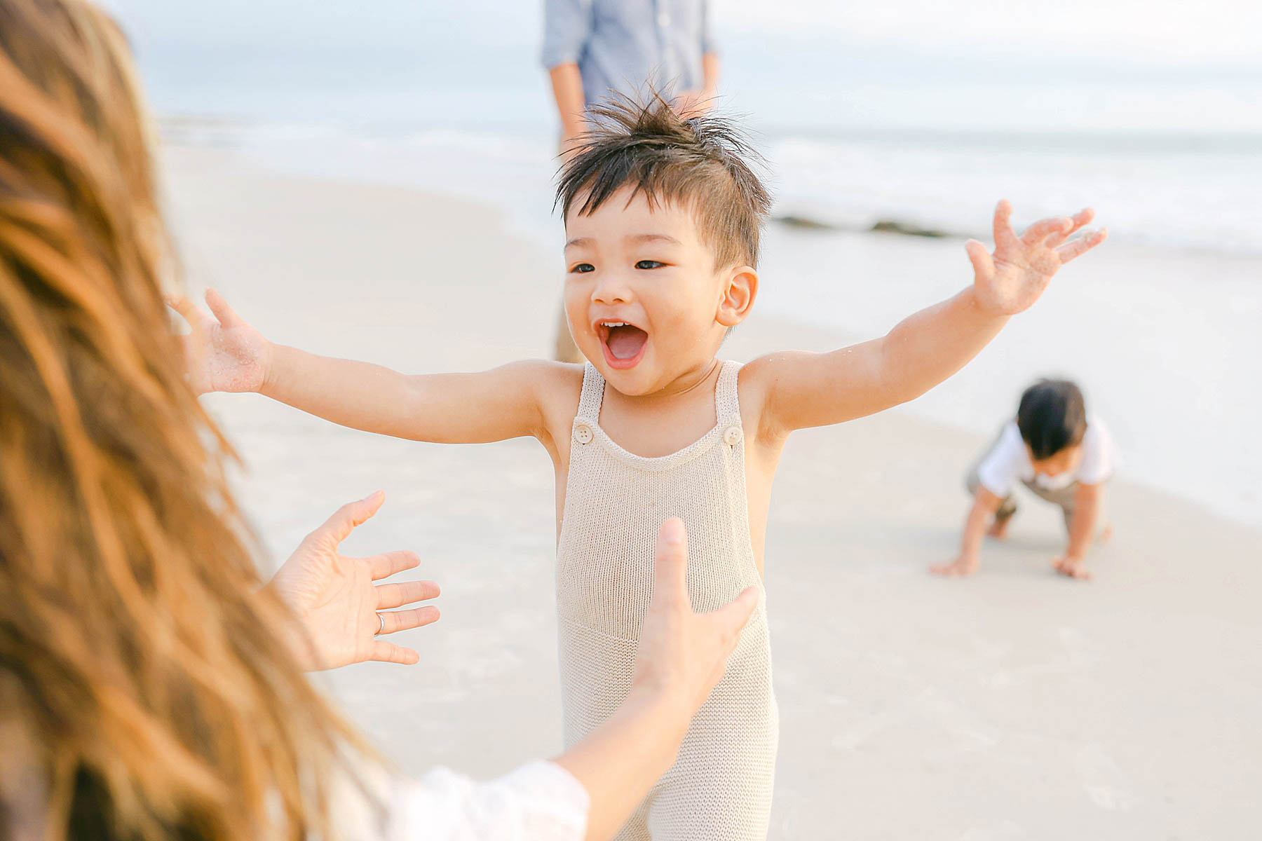little boy running to hug mom on the beach wearing overalls