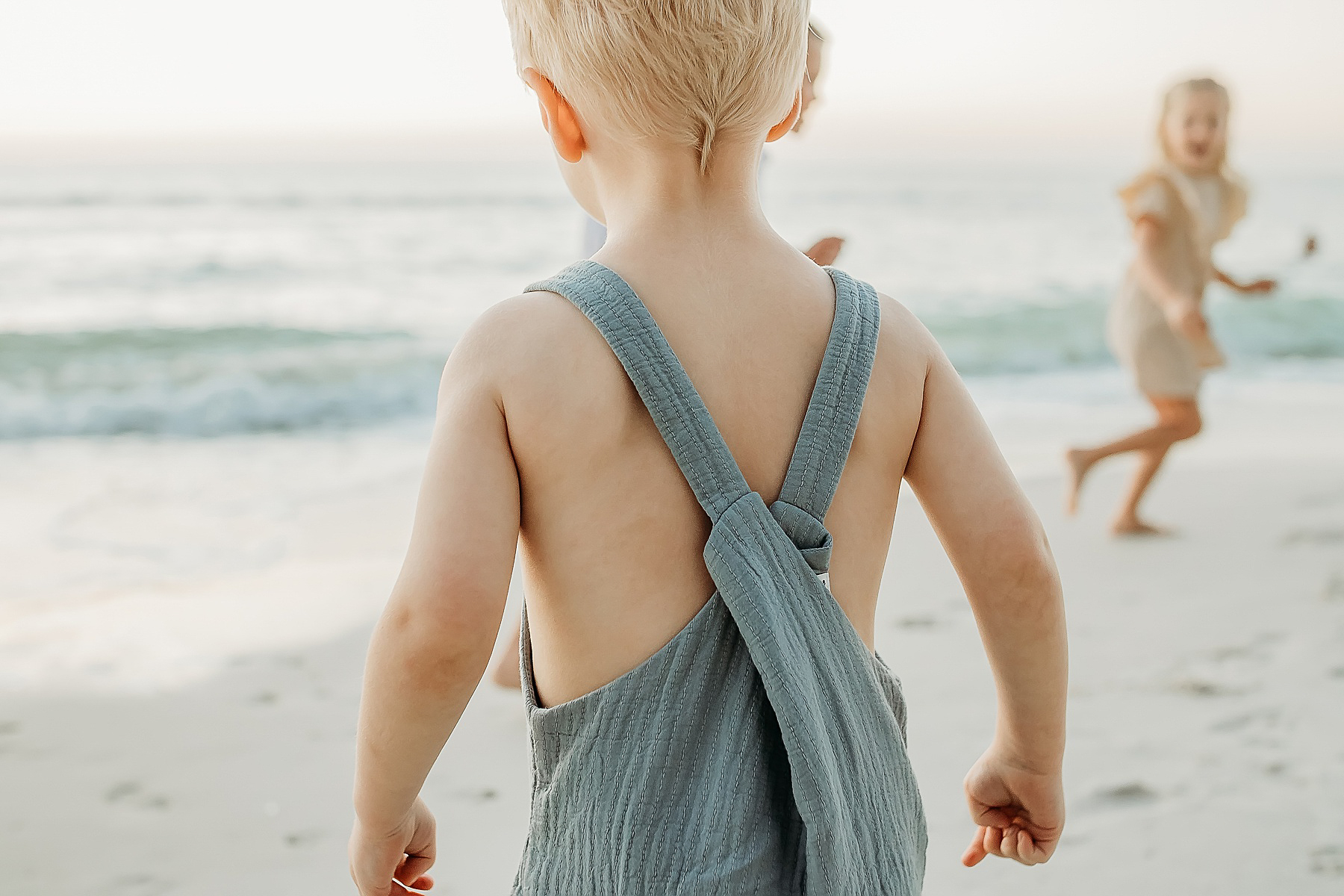 back of little boy on the beach at sunset wearing sage green overalls