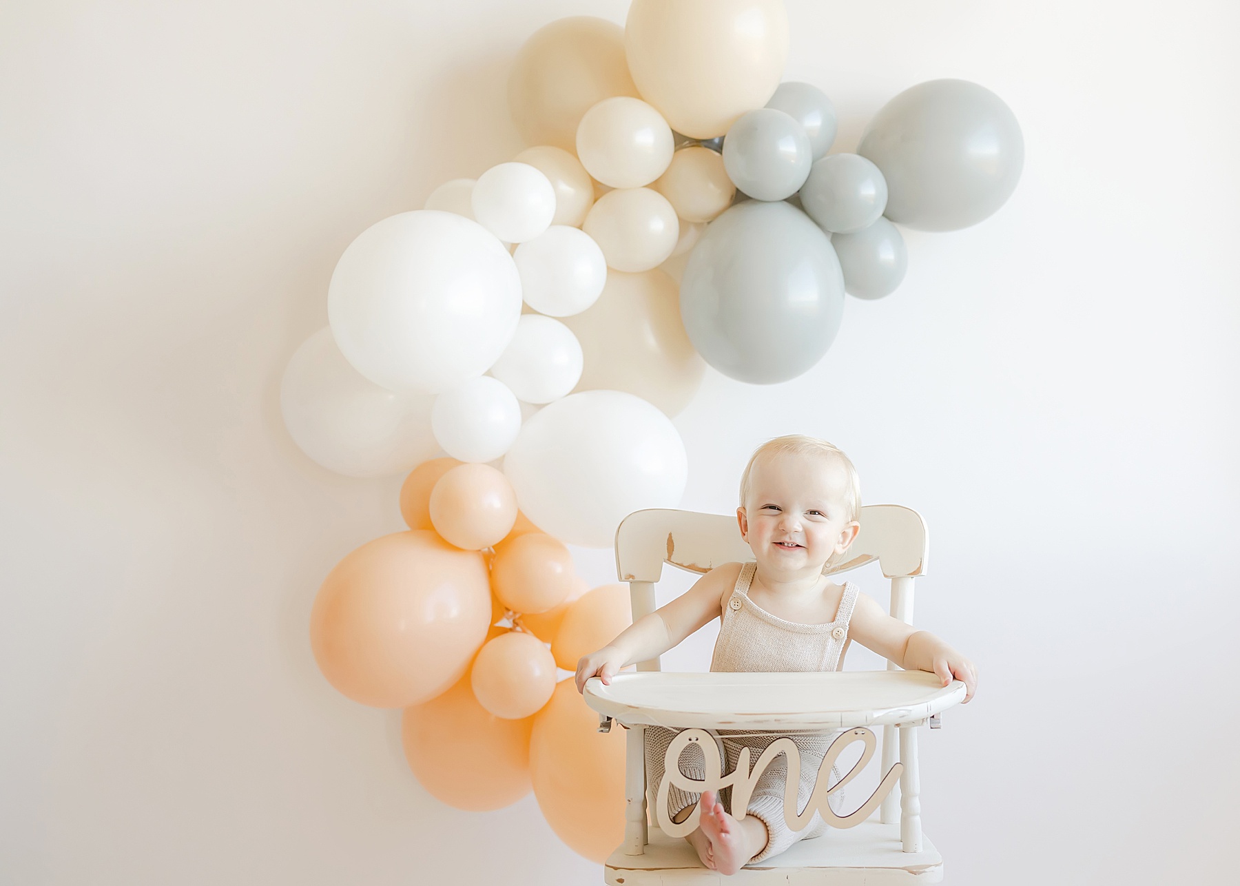 baby boy sitting in white high chair with neutral balloons behind him