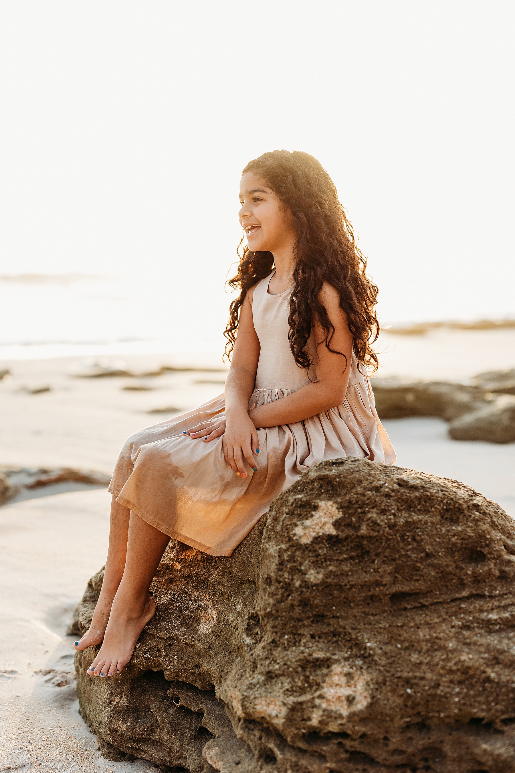 little girl sitting on a rock at the beach at sunrise wearing brown dress holding a stuffed animal fox