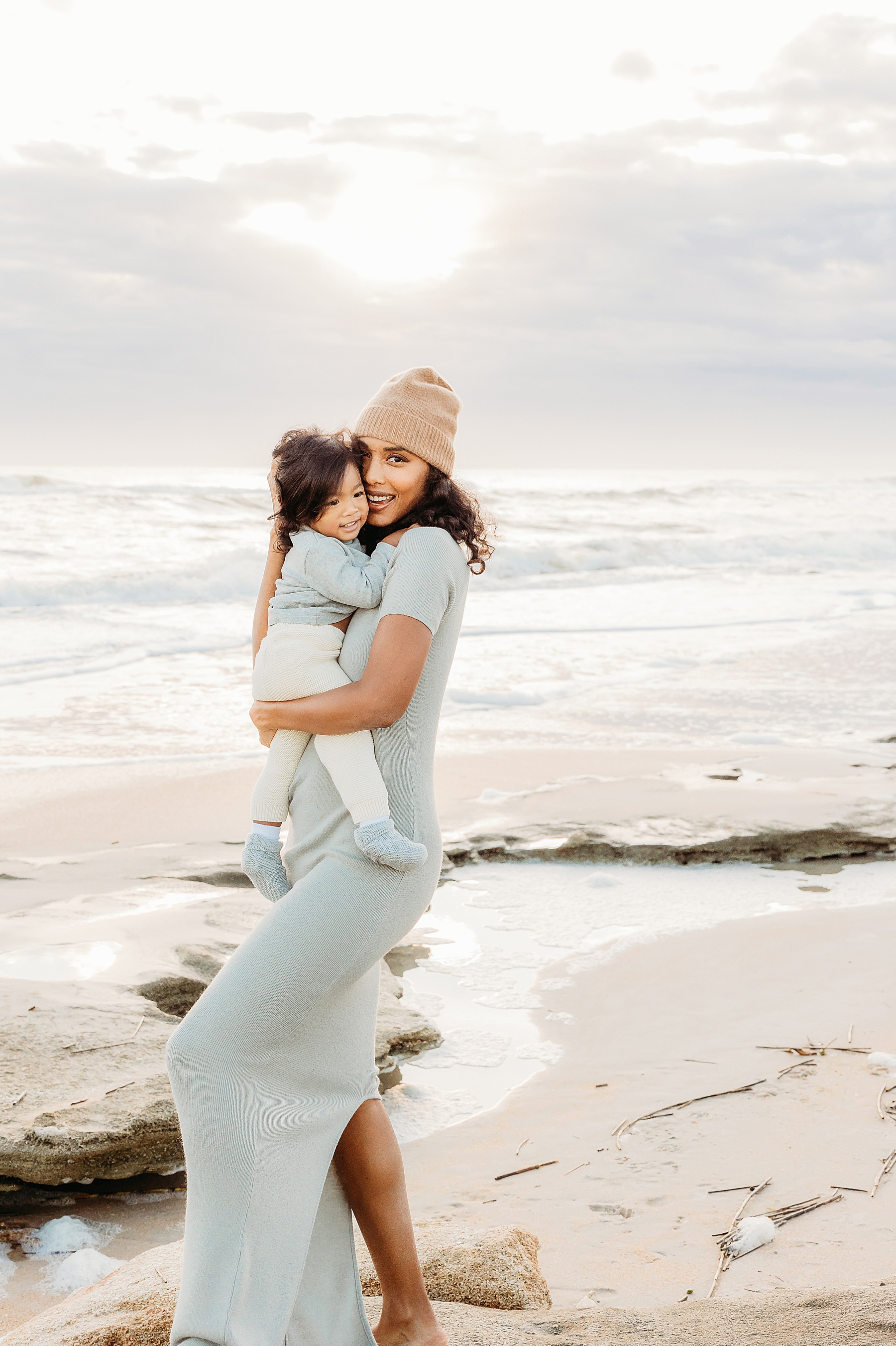 woman holding baby boy on the beach at sunrise wearing tan knit hat