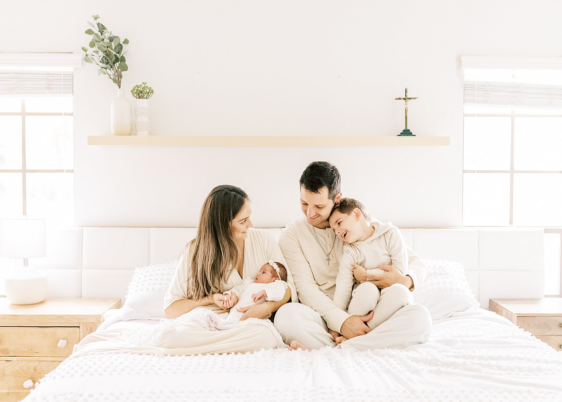 family dressed in tan and white sitting on bed with white comforter smiling at newborn baby