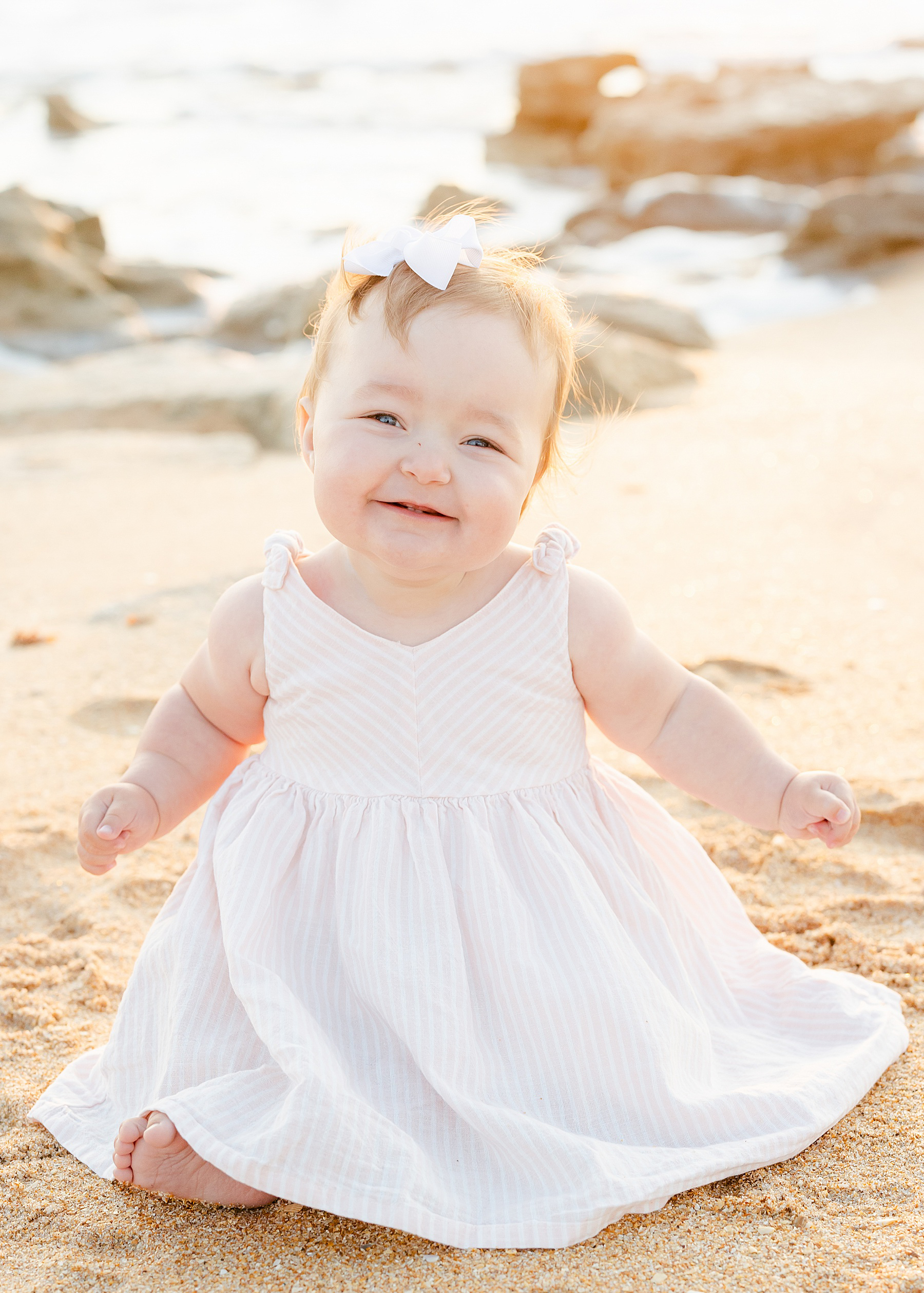 baby girl in pink and white striped dress sitting on the sand on the beach smiling
