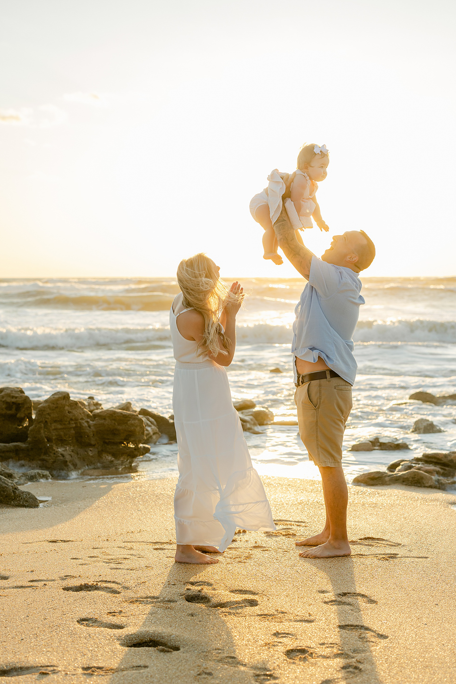 man wearing light blue shirt holding baby girl at sunrise on the beach with woman standing next to him