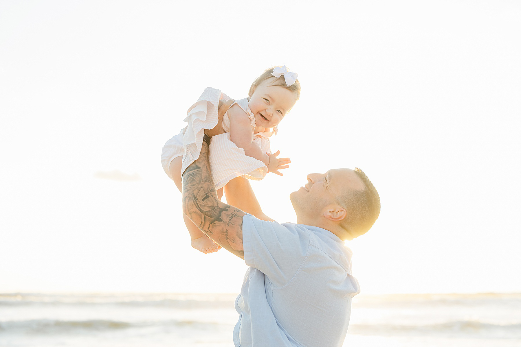 man in blue shirt holding baby girl with pink dress on the beach at sunrise smiling